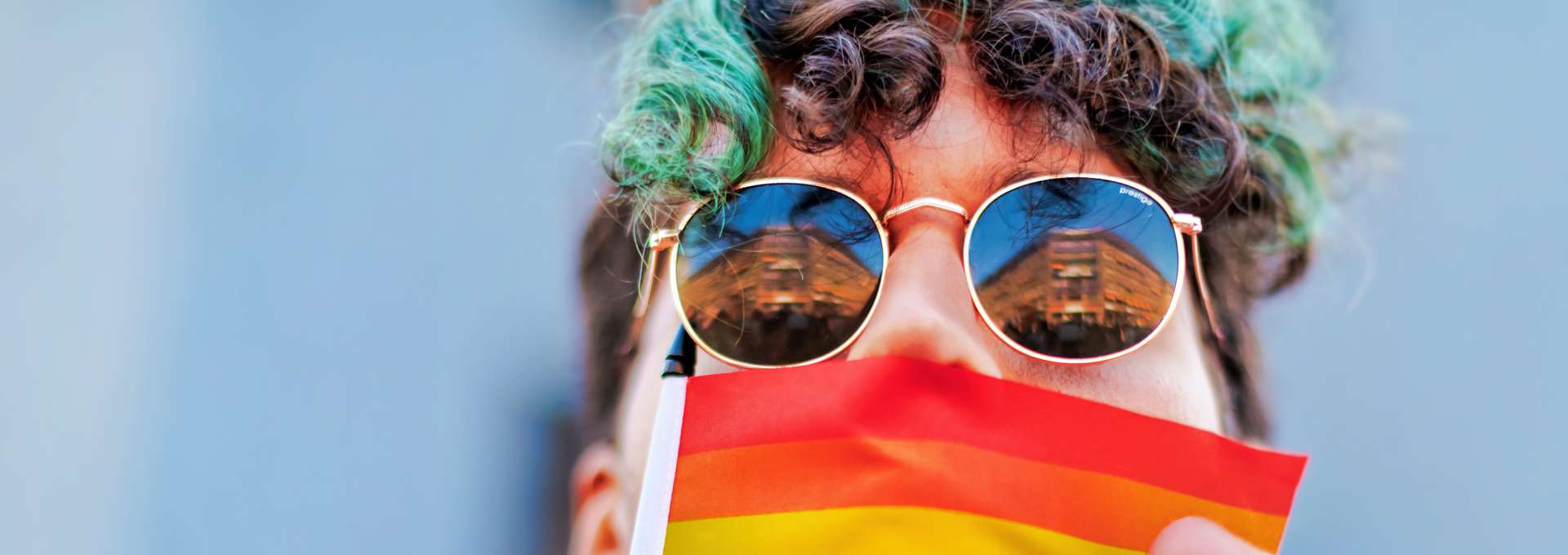 Person med lockigt brunt och grönt hår och solglasögon kysser flagga i pridefärger