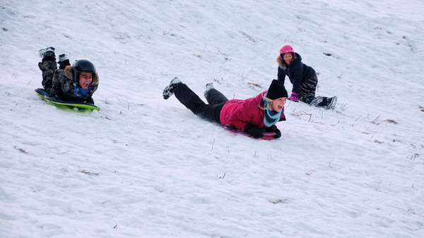 A boy and a woman are sledding on their stomachs down a hill. A girl is kneeling in the background, watching.